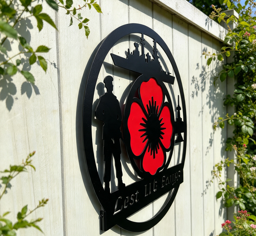 A black metal wall sign featuring a red poppy flower, soldier silhouette, and ship silhouette, mounted on a white wooden fence with green foliage in the foreground.