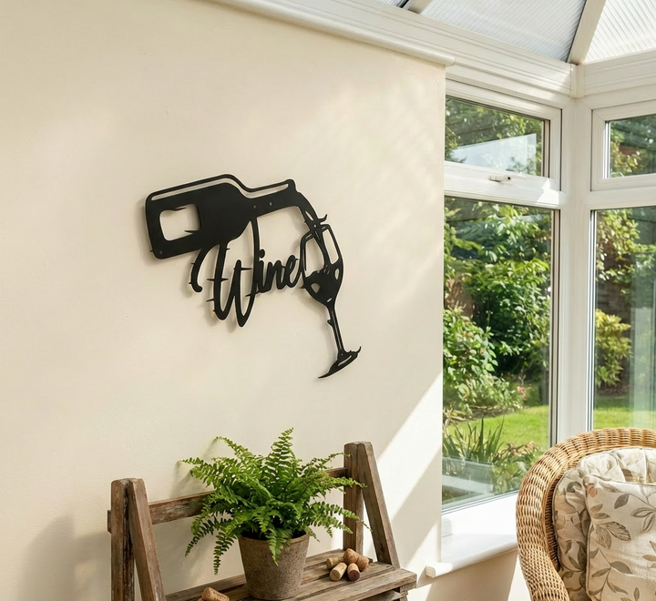 Matte black metal wall art depicting a wine bottle pouring into a glass with the word "Wine" in script, set in a conservatory setting
